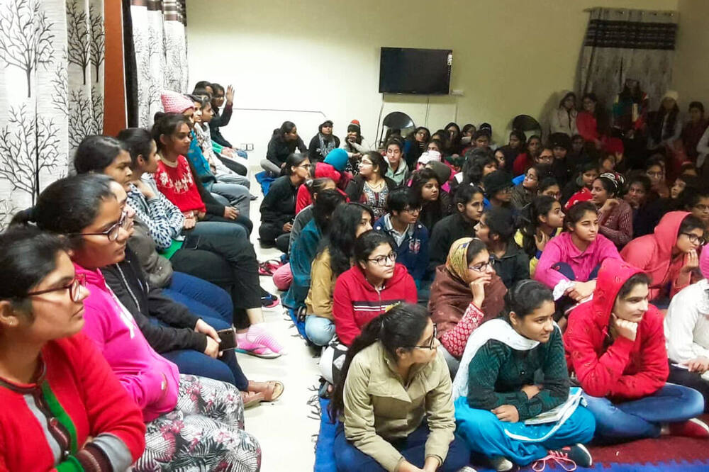 Informal indoor session, large group of young women seated on the floor, likely a HAU hostel common room